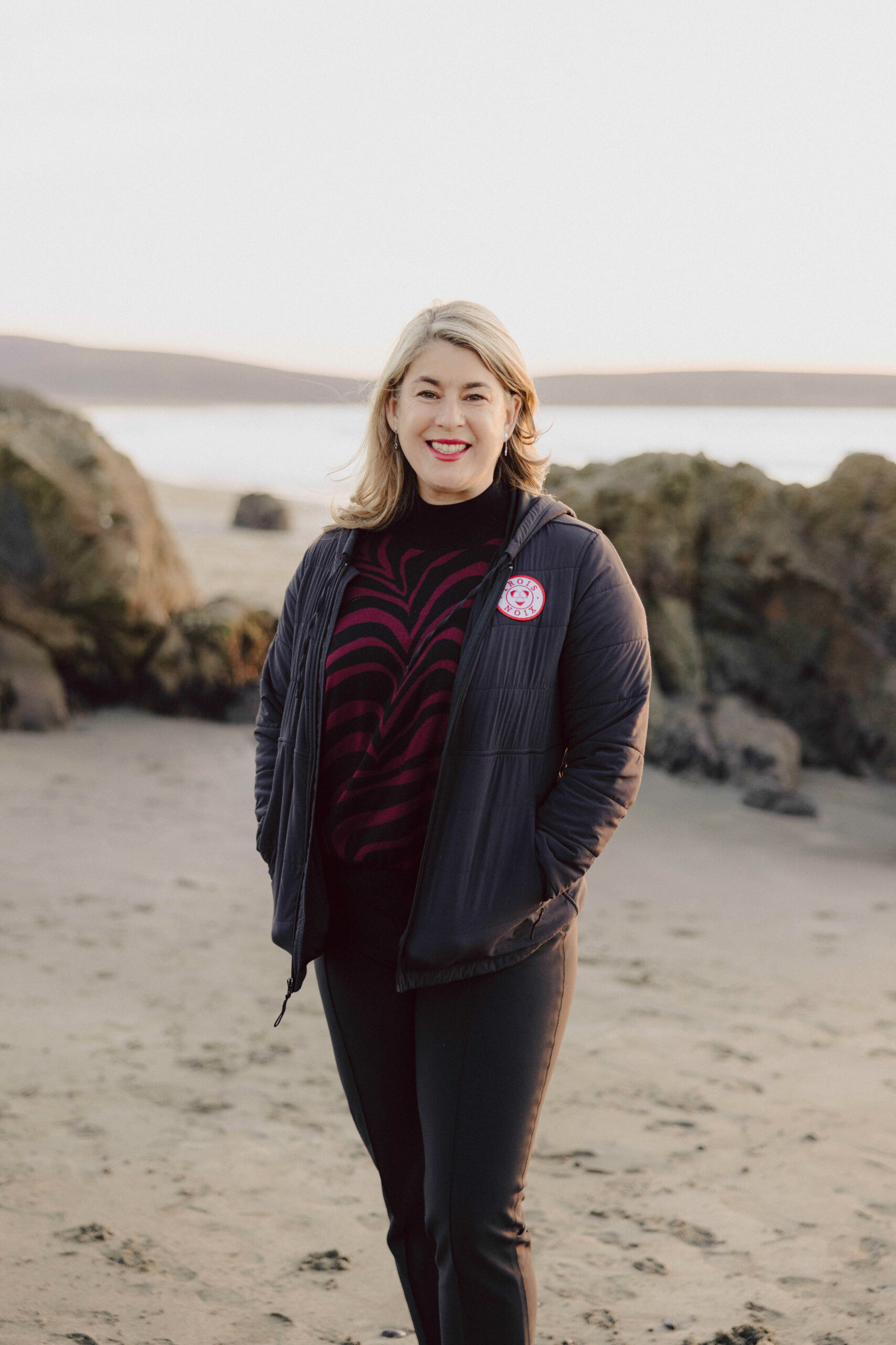 Trois Noix Founder Jaime Araujo in black jeans, a black jacket with a red and white Trois Noix patch, and a black and red sweater. She's smiling and standing with her hands in her pockets in front of rocks on Dillon Beach, CA.
