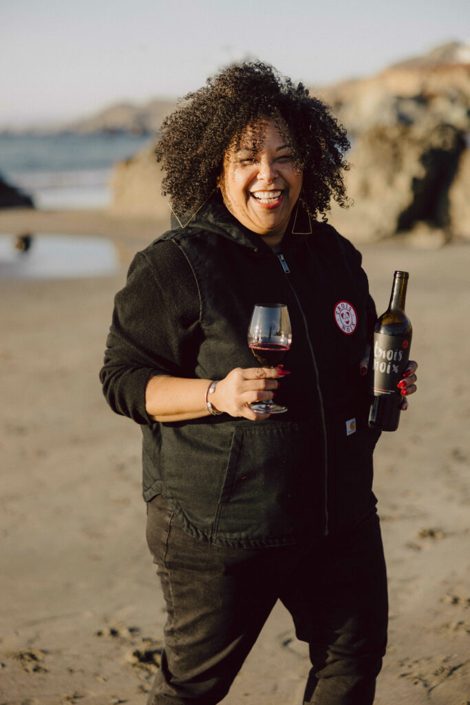 Suhayl Ramirez standing on a beach smiling with a bottle and glass of cabernet sauvignon in her hand