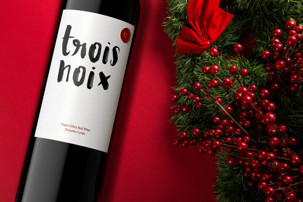 A bottle of Trois Noix Noisette wine against a red background with a green holiday wreath with red berries and bow is a perfect corporate gift.